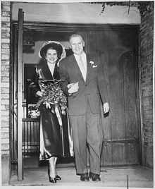 A man in a suit leads a flower-carrying woman by the hand, walking out of a chapel.